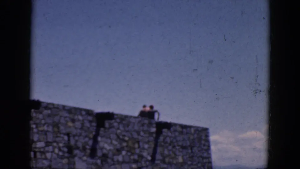 1964 Charlestown, North Carolina home-movie frame with two distant people resting on a stone wall beneath the sky.