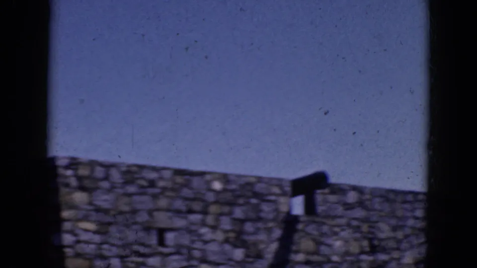 1964 8mm frame from the same stone-wall scene, showing the wall, sky, and dark frame edges with people not clearly resolved.