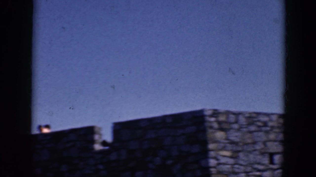 Stone wall and red roof under blue sky, framed by dark 8mm film edges.