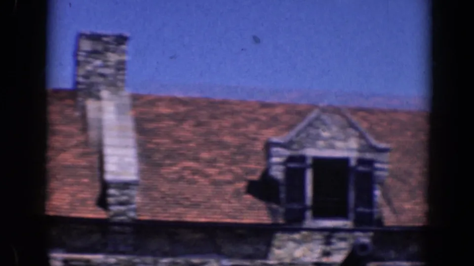 1964 8mm frame showing a chimney and dormer on a red-tiled roof, with stone trim and blue sky.