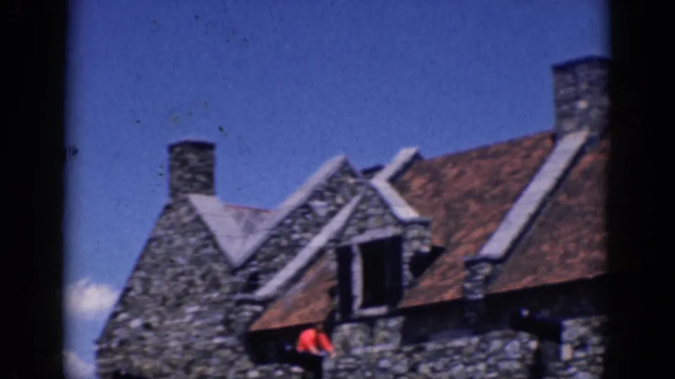 1964 home-movie view of a gabled stone building with red-tiled roof and a distant person in red near the lower edge.