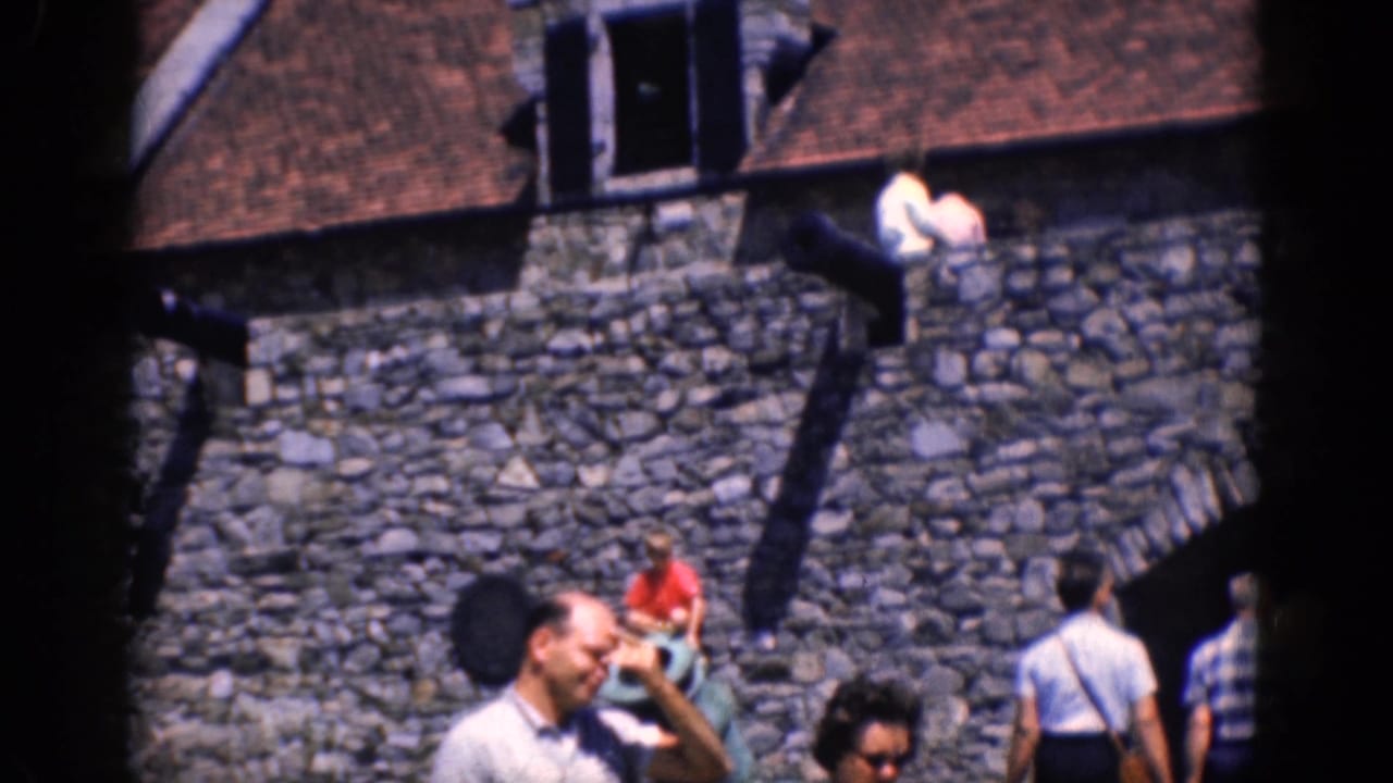 1964 home-movie frame of tourists at a stone fortress in Charlestown, North Carolina, with ramparts, steps, and a red-shirted visitor.
