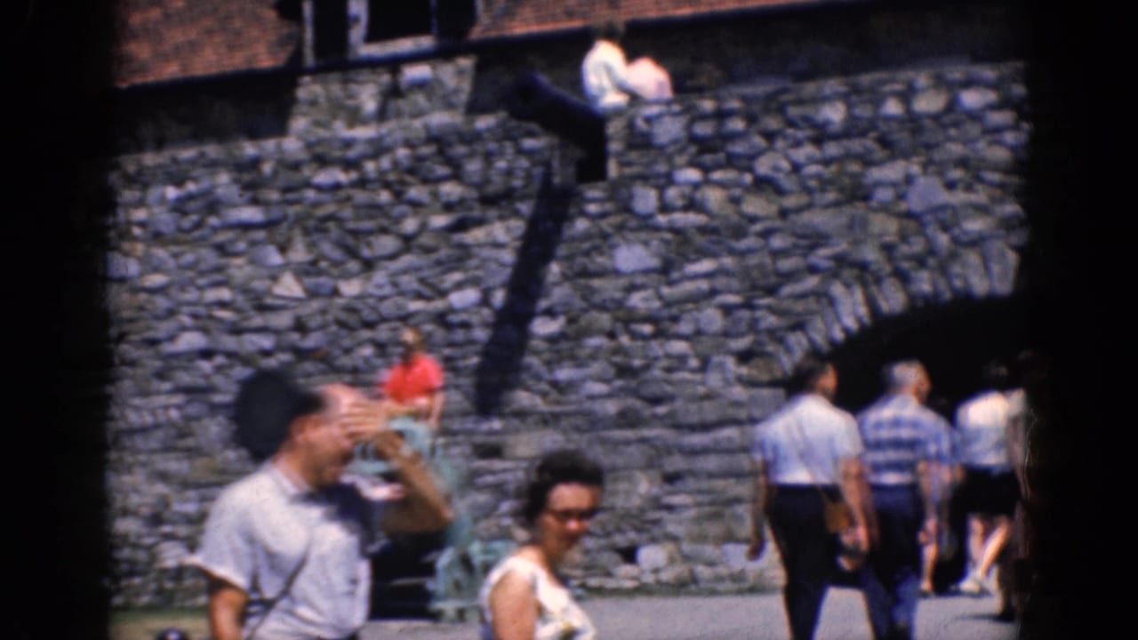 1964 8mm frame showing tourists at the Charlestown stone fortress, with foreground visitors and rampart stairs in sunlight.