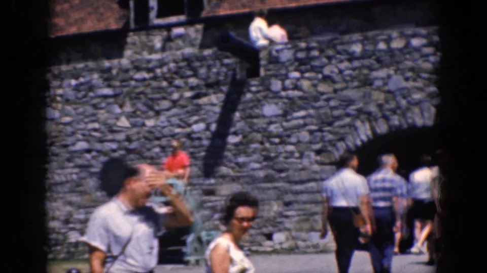 1964 8mm frame showing tourists at the Charlestown stone fortress, with foreground visitors and rampart stairs in sunlight.