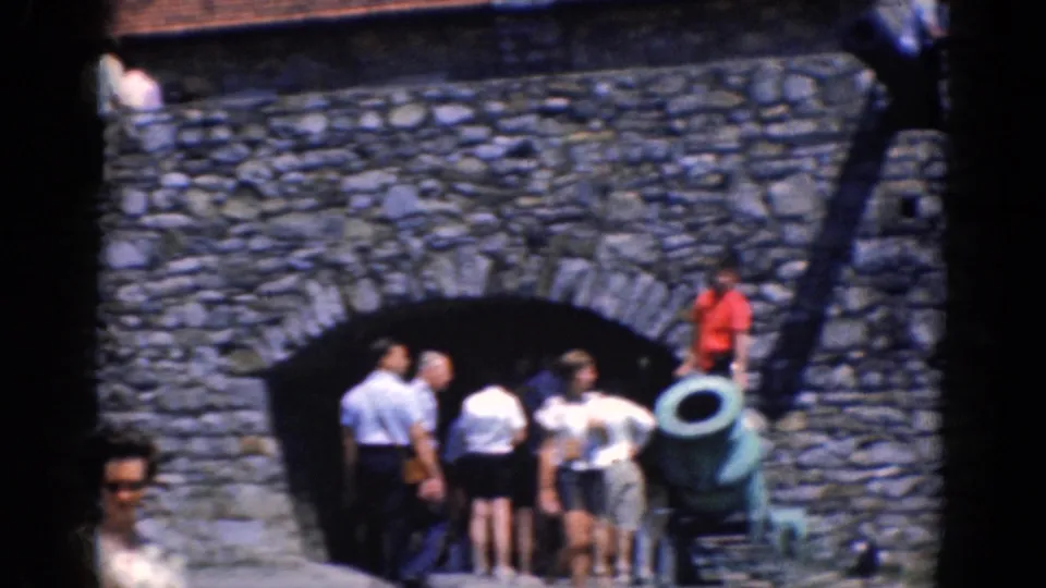 Final 1964 home-movie frame of visitors clustered around a cannon before a stone arch at the Charlestown historic site.
