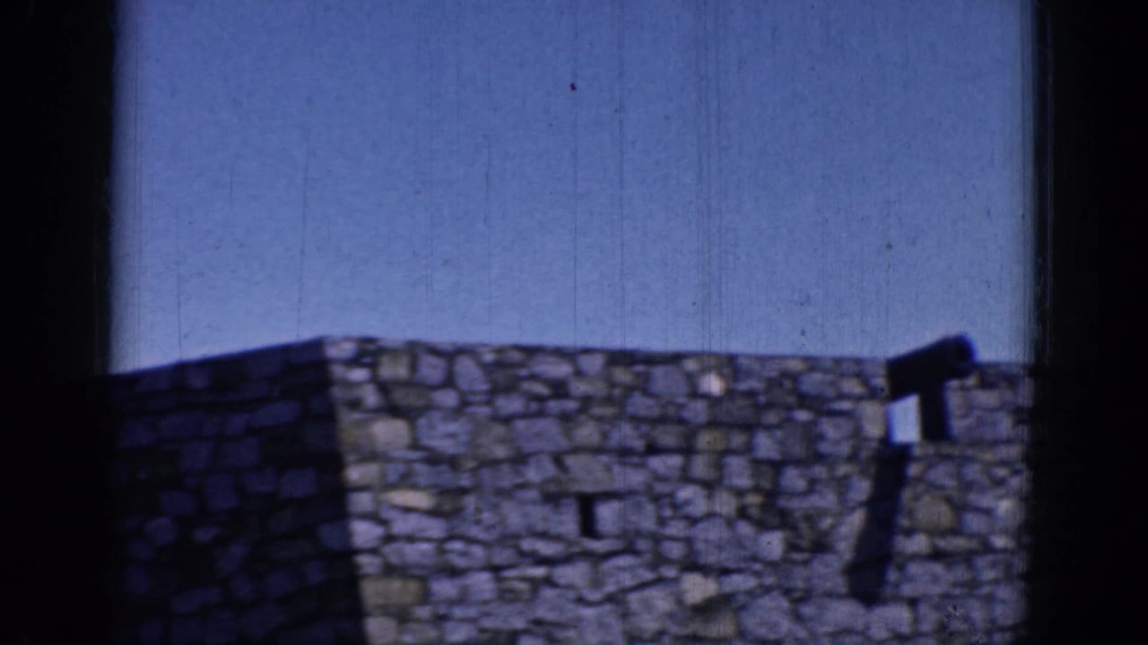 1964 8mm frame from Charlestown, North Carolina showing a rough stone wall and crenellated top beneath clear blue sky.