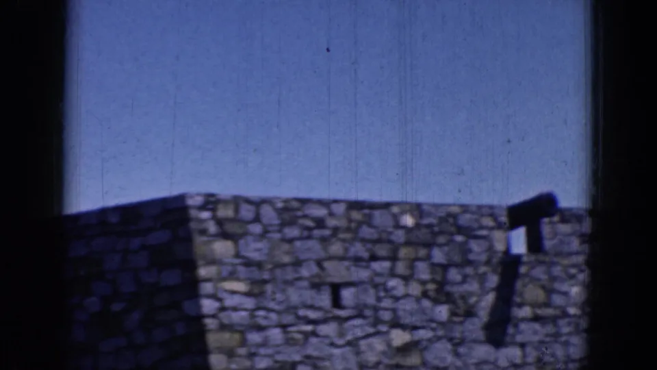 1964 8mm frame from Charlestown, North Carolina showing a rough stone wall and crenellated top beneath clear blue sky.