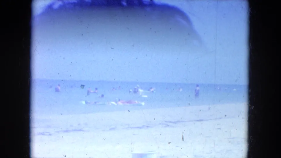 1964 Clearwater, Florida beach scene with distant swimmers in shallow water, pale sand, blue sky, and visible film gate edges.