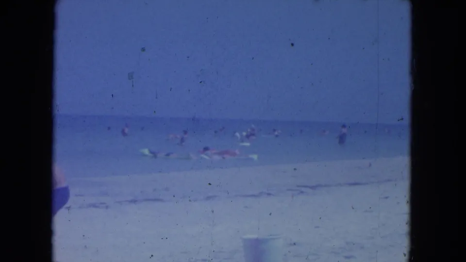 Clearwater beach footage from 1964 showing distant swimmers, floats, pale surf, and a partial foreground figure near the film edge.