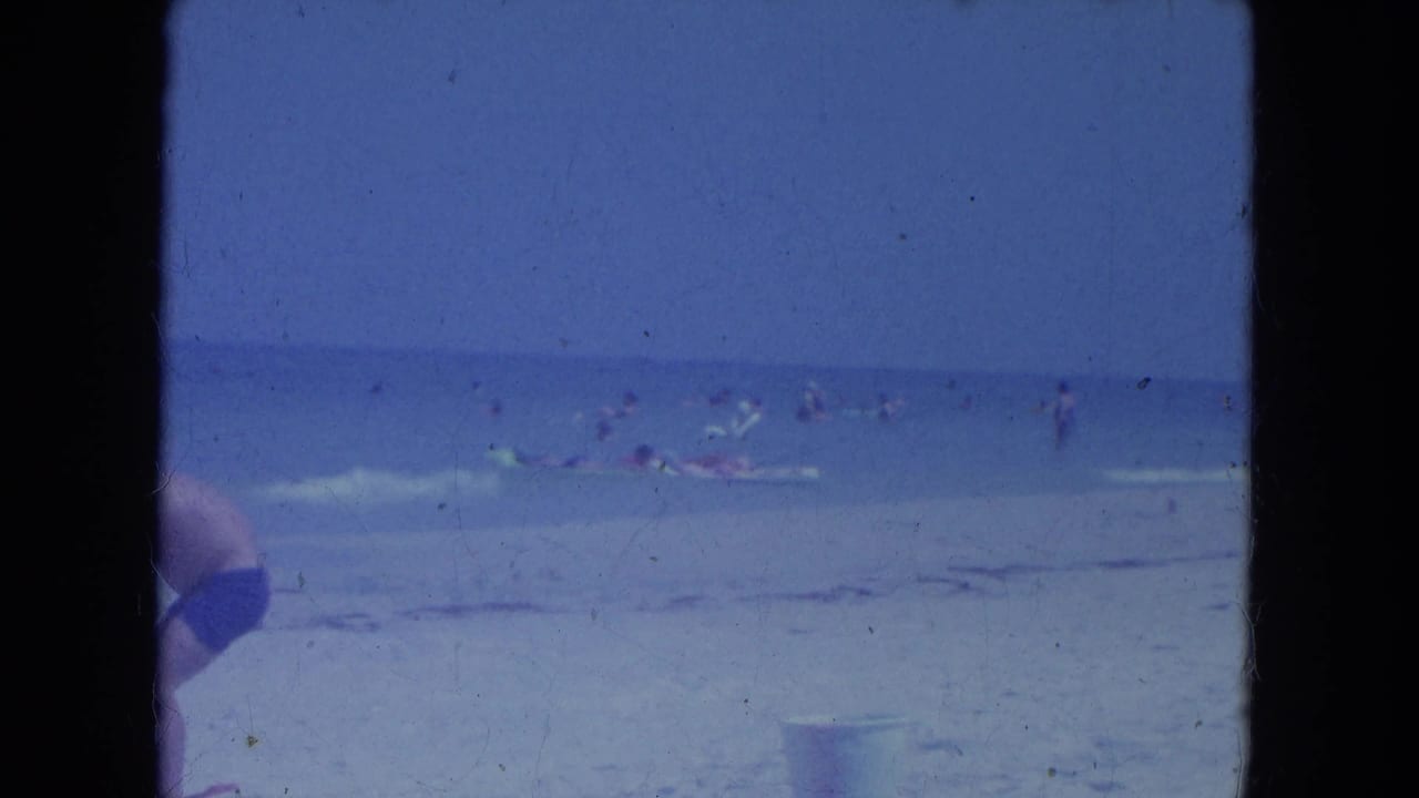 Blue-shifted 8mm view of Clearwater beach in 1964, with swimmers offshore, sand in front, and a partial swimsuit figure at left.