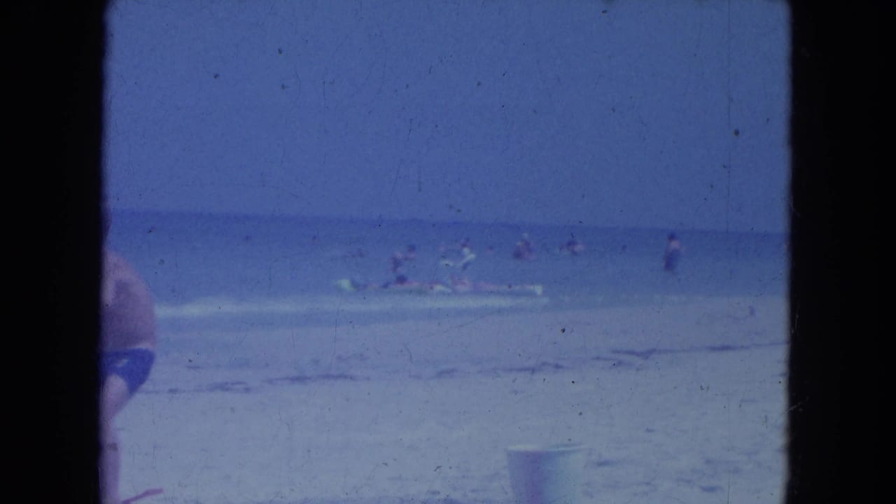 1964 Clearwater beach frame with swimmers, floats, a partial foreground swimsuit figure, and a small bucket-like object on the sand.