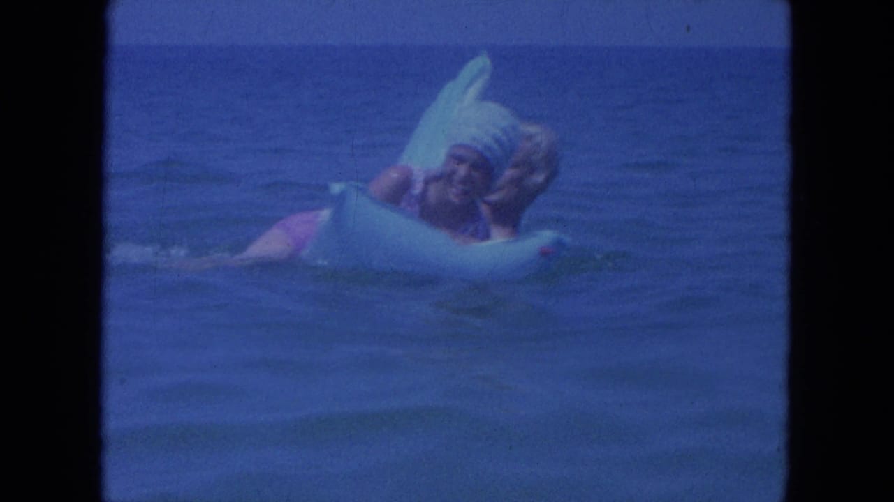 Two adults laugh together on a light blue inflatable ring in ocean water.