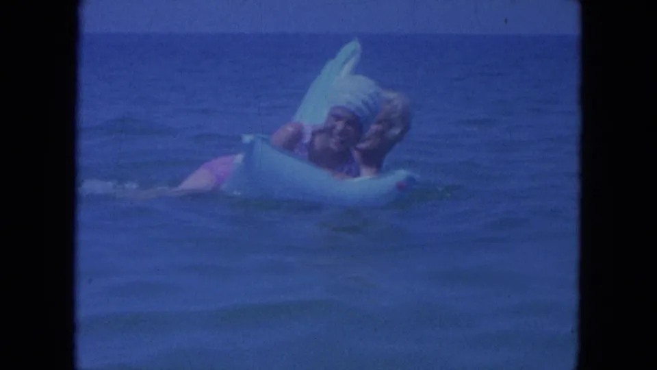In 1964 Clearwater, Florida footage, a woman in a swim cap smiles on a light blue float while a man holds close in the ocean.