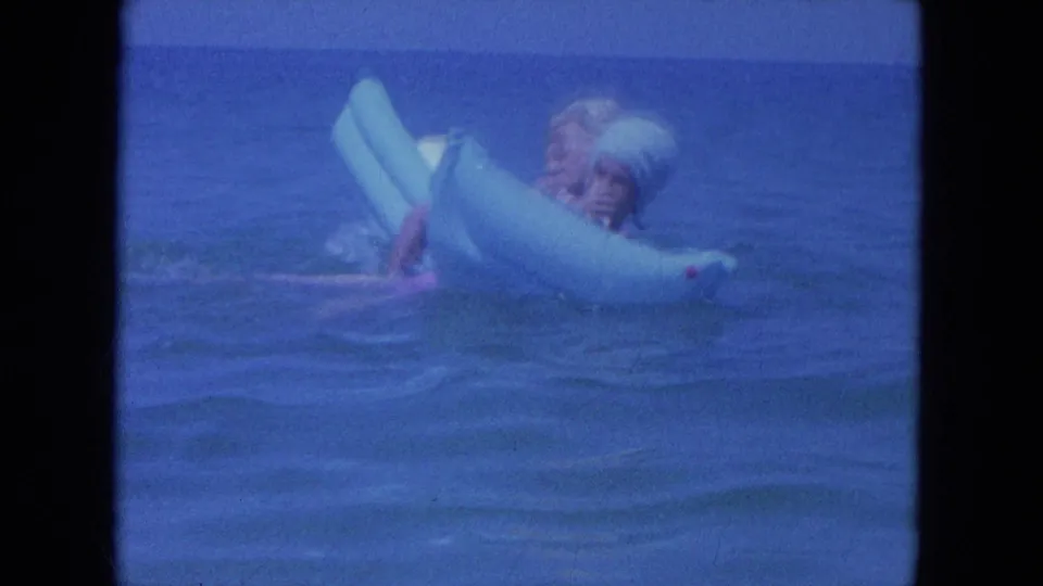 A couple appears side-on in the 1964 Clearwater ocean scene, balancing together on a pale blue inflatable ring.