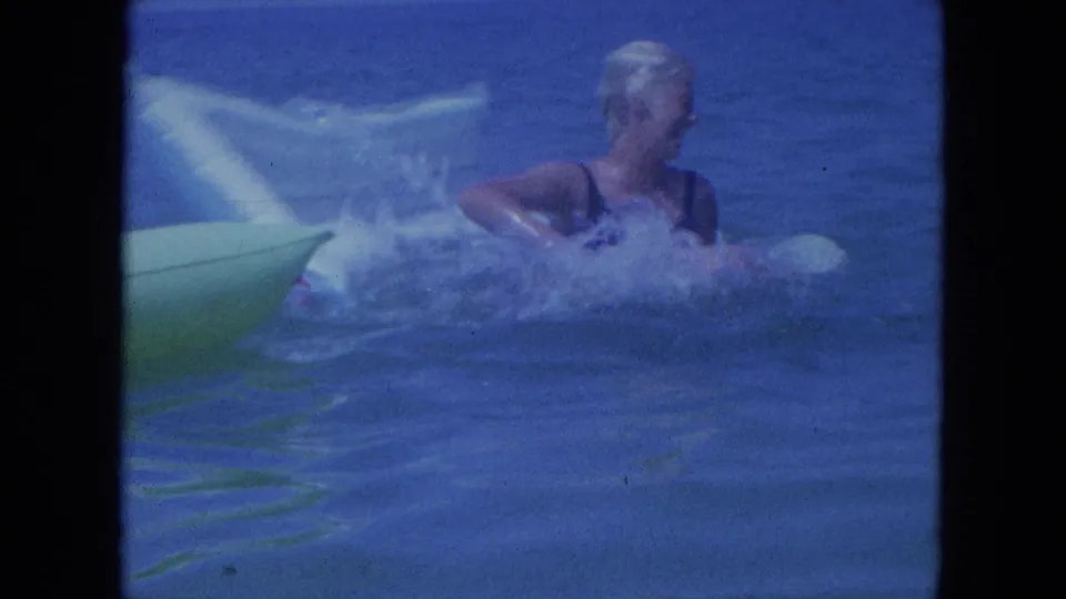 1964 Clearwater home-movie frame showing a woman in a swim cap splashing near a green inflatable float in blue water.