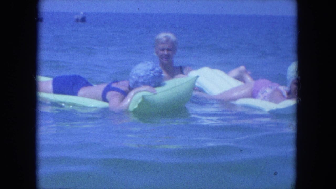1964 Clearwater home-movie frame showing women lounging on green and white floats while another sits upright in the water.