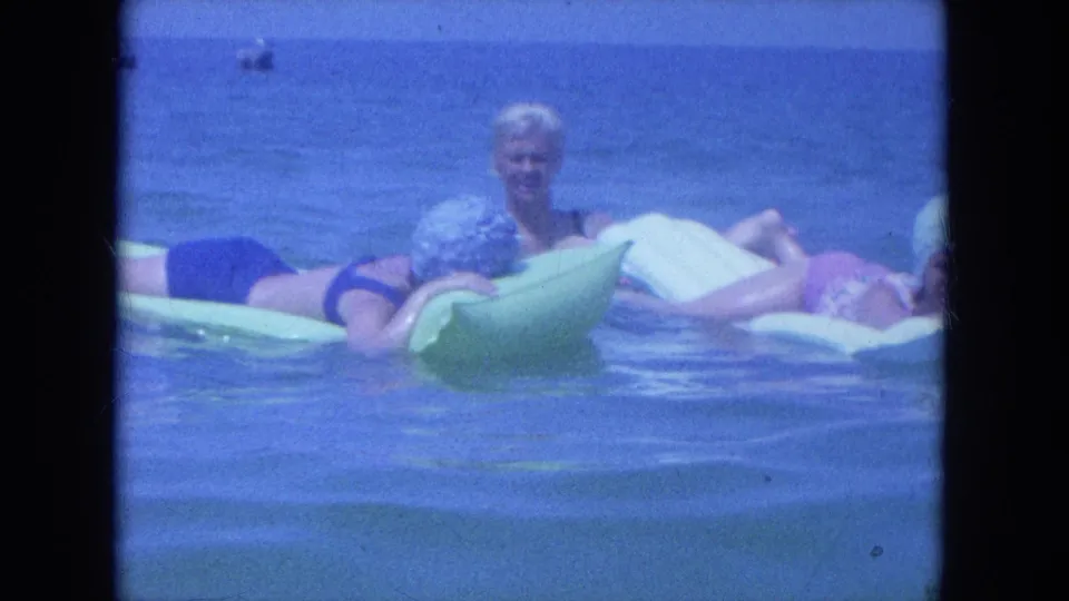 1964 Clearwater home-movie frame showing women lounging on green and white floats while another sits upright in the water.