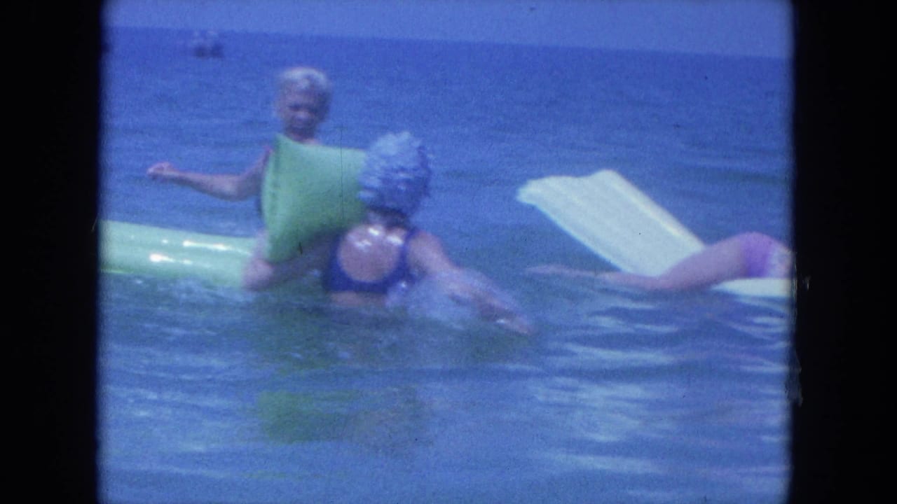 A woman in a blue swim cap moves through the water near another woman resting against a green inflatable raft.