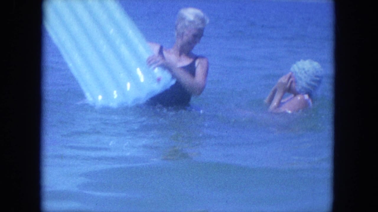 In 1964 Clearwater beach footage, two women stand in blue ocean water while handling a pale inflatable mat.