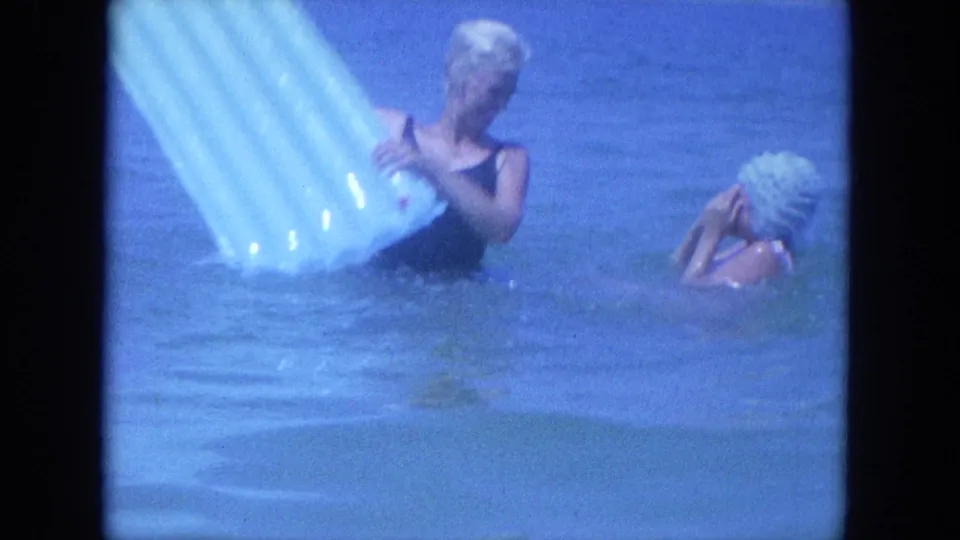 In 1964 Clearwater beach footage, two women stand in blue ocean water while handling a pale inflatable mat.