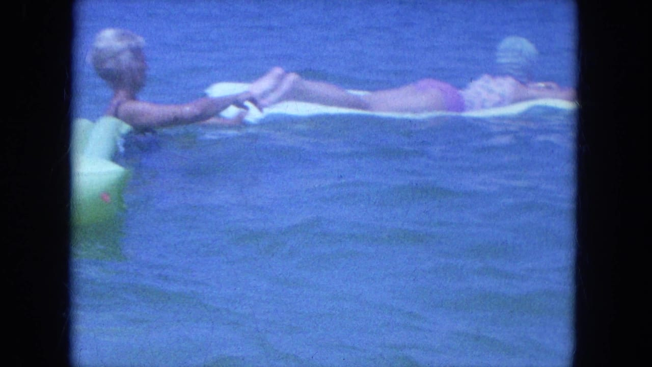 A woman floats on a pale inflatable mat as another woman holds the mat in 1964 Clearwater ocean water.