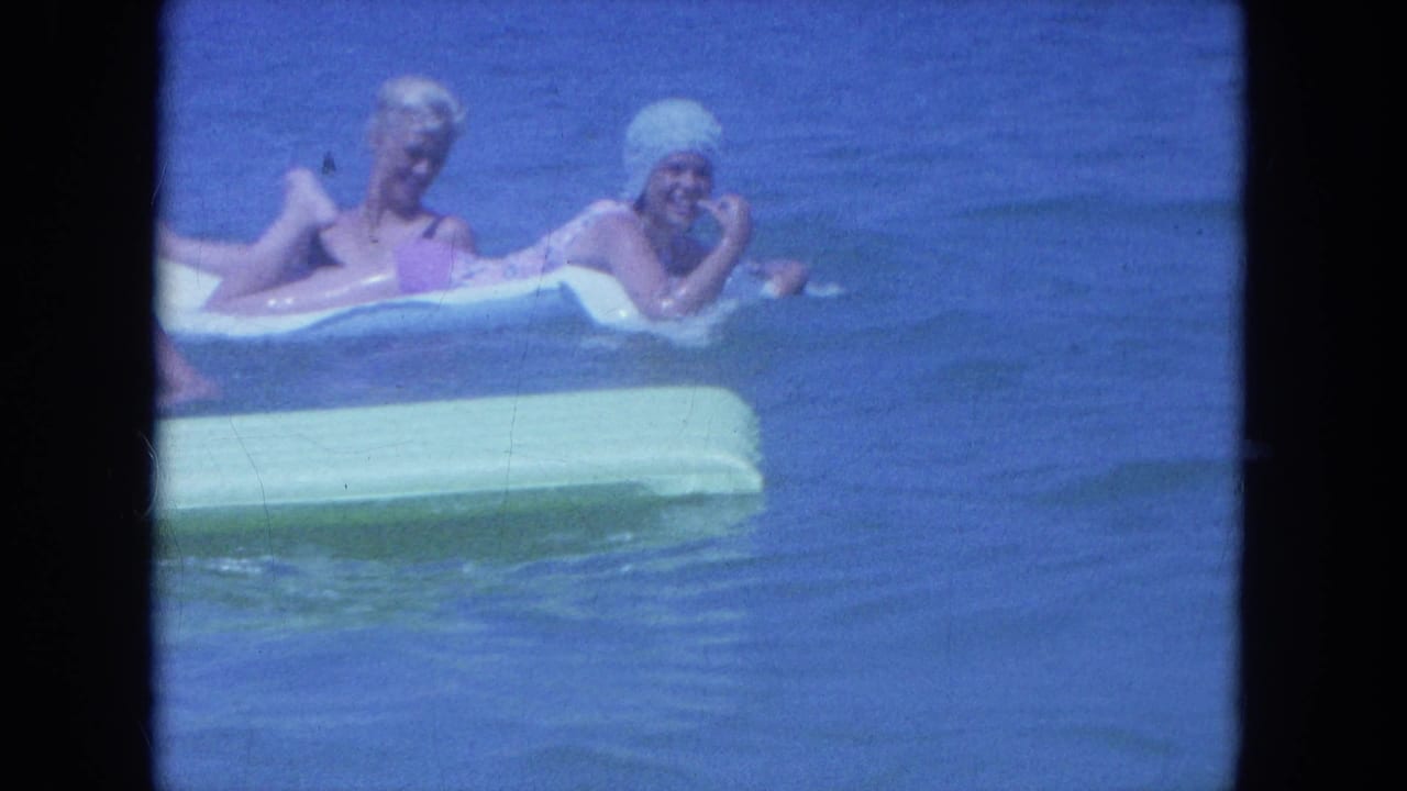 Two adult women in swim caps and bathing suits float close to the camera on rafts in 1964 Clearwater beach footage.