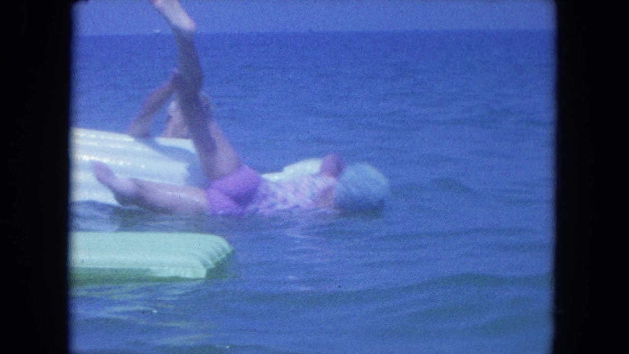 An adult woman in a swim cap splashes backward from a raft in blue ocean water during the Clearwater home movie clip.