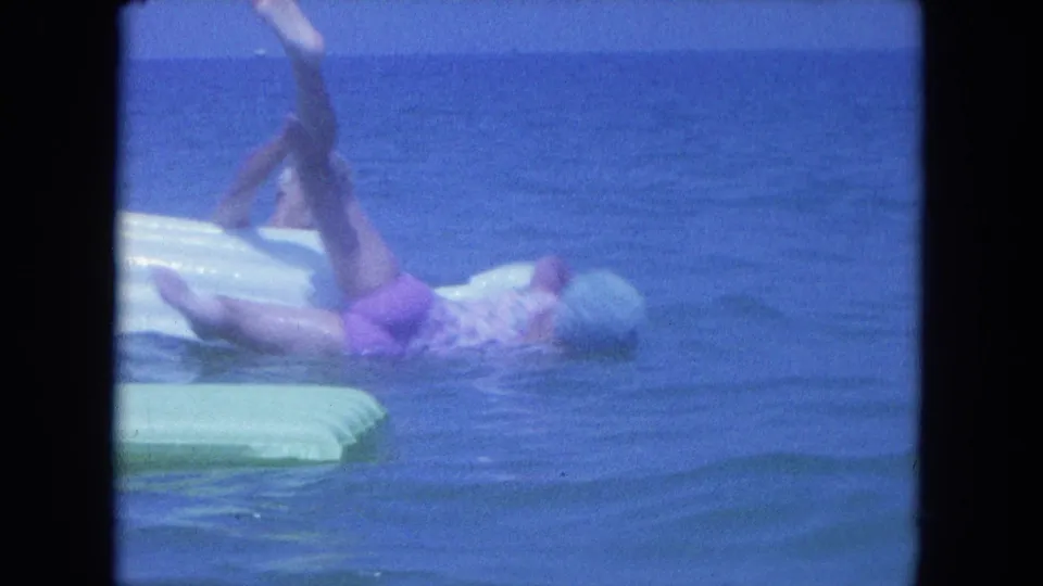 An adult woman in a swim cap splashes backward from a raft in blue ocean water during the Clearwater home movie clip.