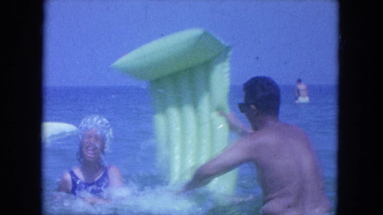 Woman in a blue swimsuit and white swim cap splashes near a man holding a green inflatable float in the ocean.