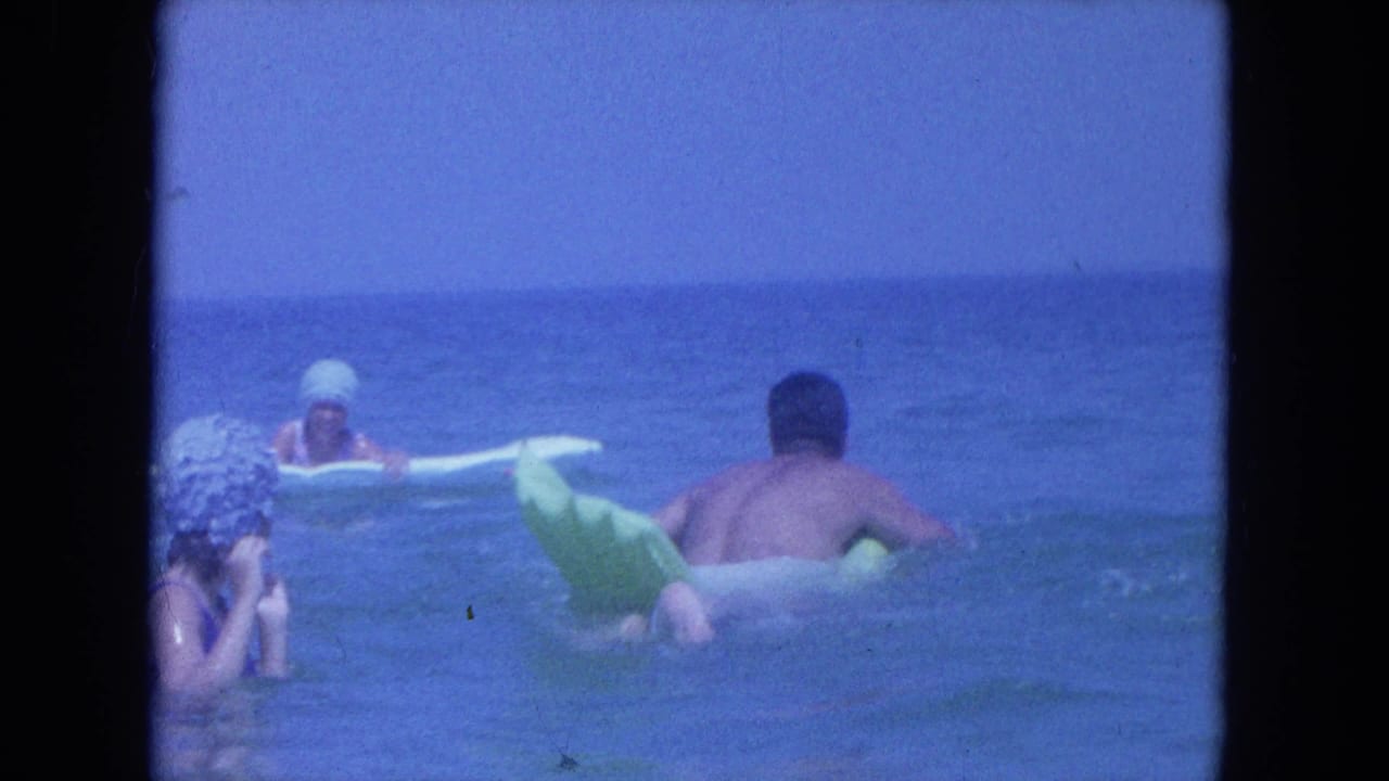 1964 Clearwater beach home-movie frame shows a woman in a white swim cap at left while a man rests on a green inflatable float in shallow surf.