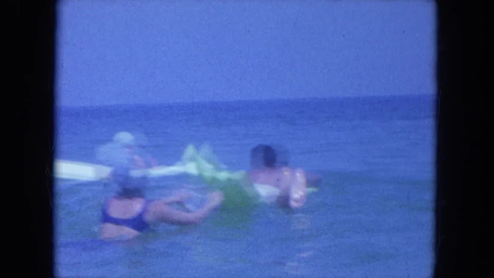 Clearwater beach footage from 1964 shows adults in the surf around a green inflatable float, with another swimmer on a white float in the background.