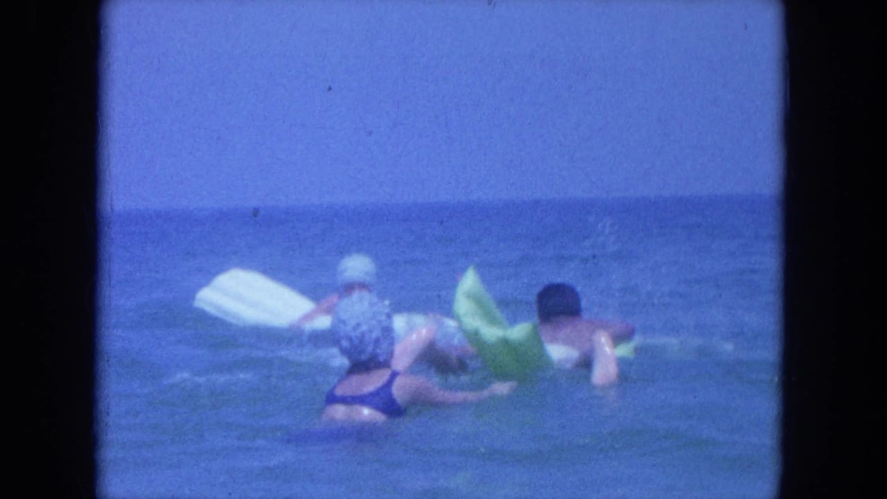 1964 Clearwater, Florida home-movie frame showing swimmers in the ocean with a white board and green inflatable float.