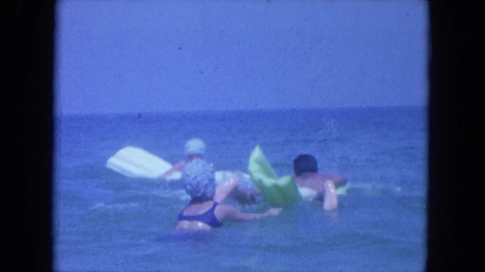 1964 Clearwater, Florida home-movie frame showing swimmers in the ocean with a white board and green inflatable float.
