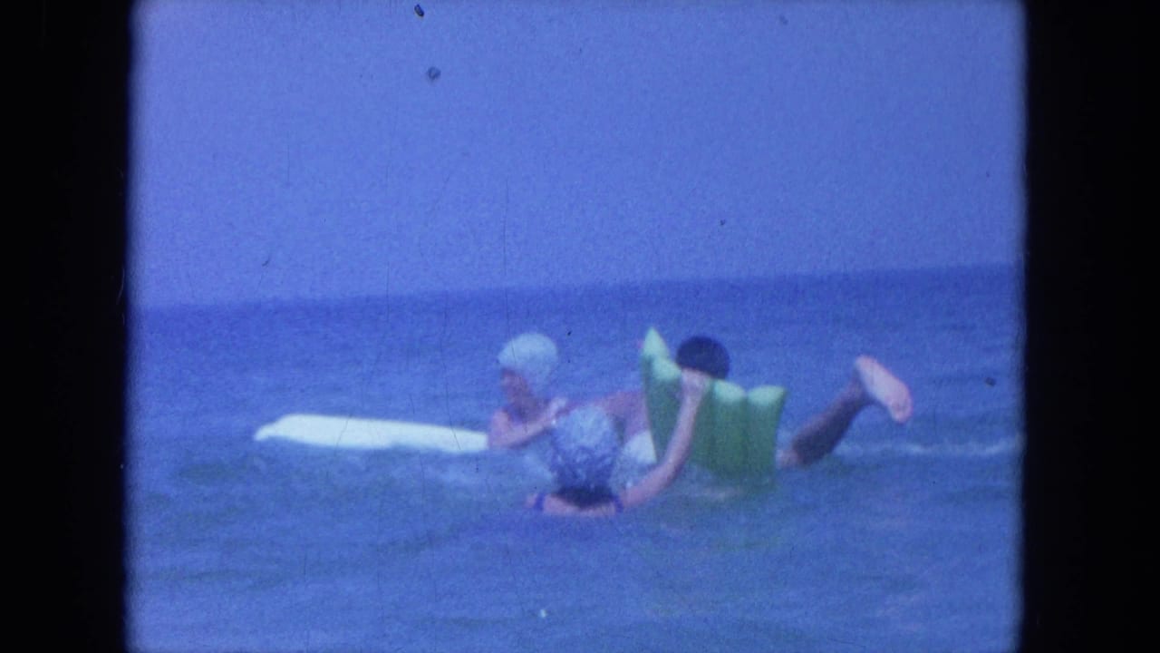 1964 Clearwater, Florida frame of a swim-capped woman and man using a green inflatable float near a white board.