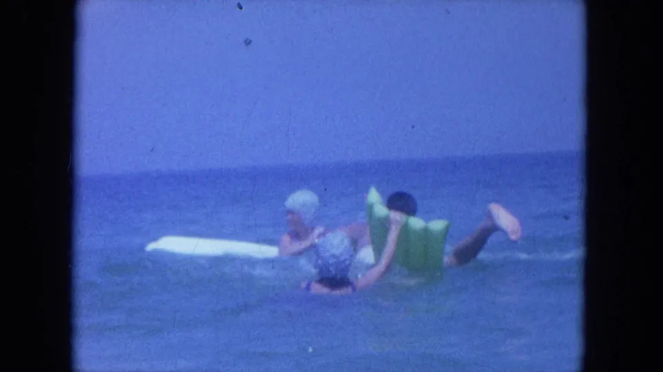 1964 Clearwater, Florida frame of a swim-capped woman and man using a green inflatable float near a white board.