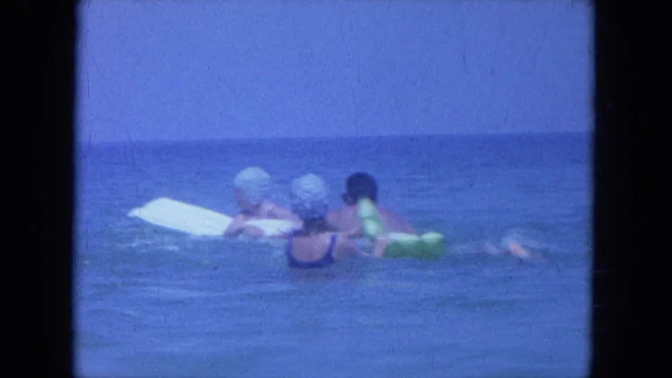 1964 Clearwater, Florida home-movie frame with three swimmers near a white board in calm open water.
