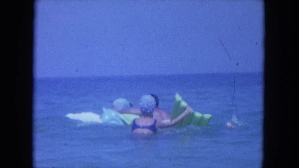 1964 Clearwater, Florida frame showing a woman in a swim cap near a green inflatable float and white board in open water.