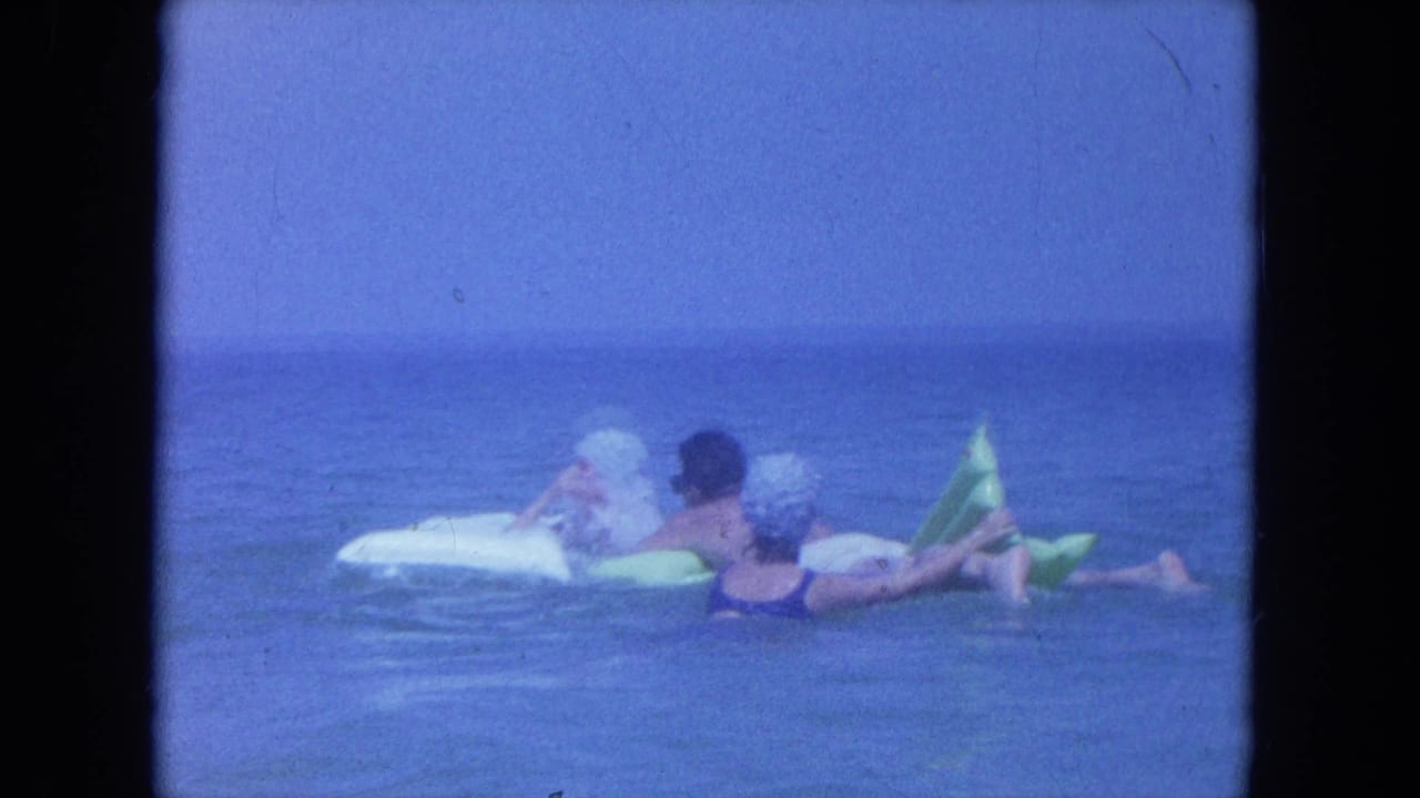 Two adults in swim caps float on inflatable rafts in calm ocean water.