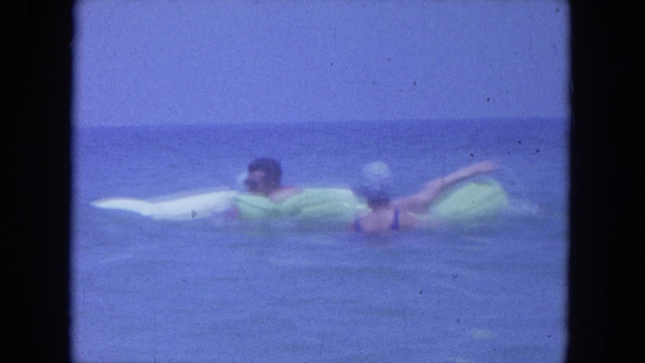 Two adults paddle on inflatable rafts in open blue water.