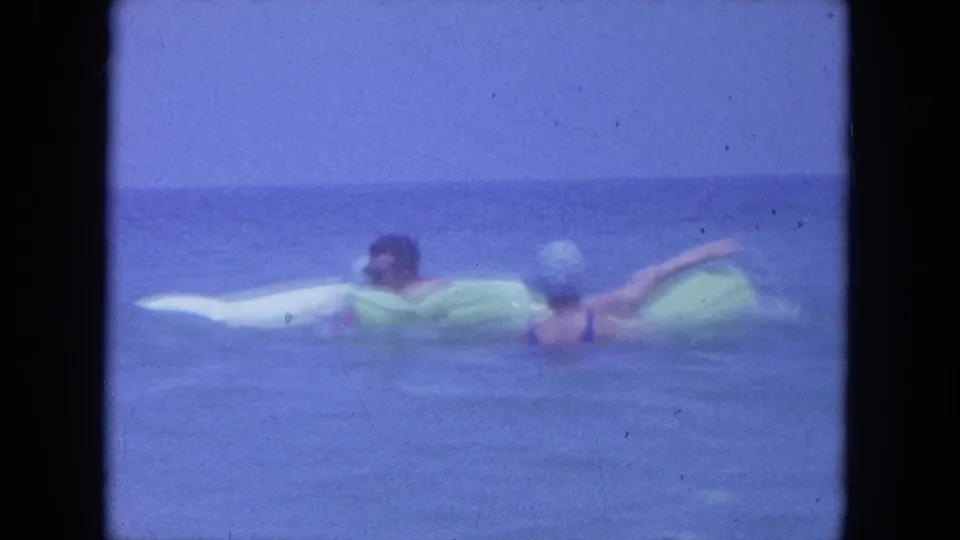 Two adults on pale green and white inflatable rafts paddle through calm ocean water in the 1964 Clearwater beach clip.
