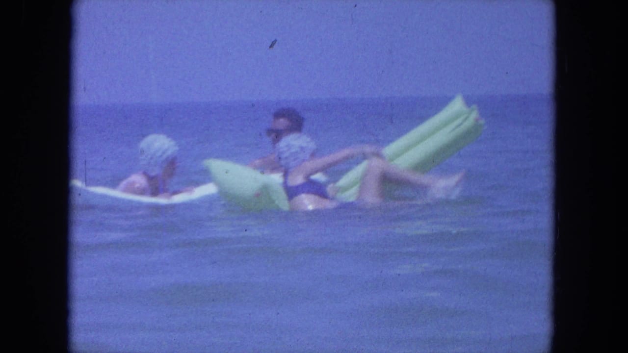 Two adults in swim caps and swimsuits recline on inflatable rafts, seen in soft 8mm color off Clearwater, Florida.