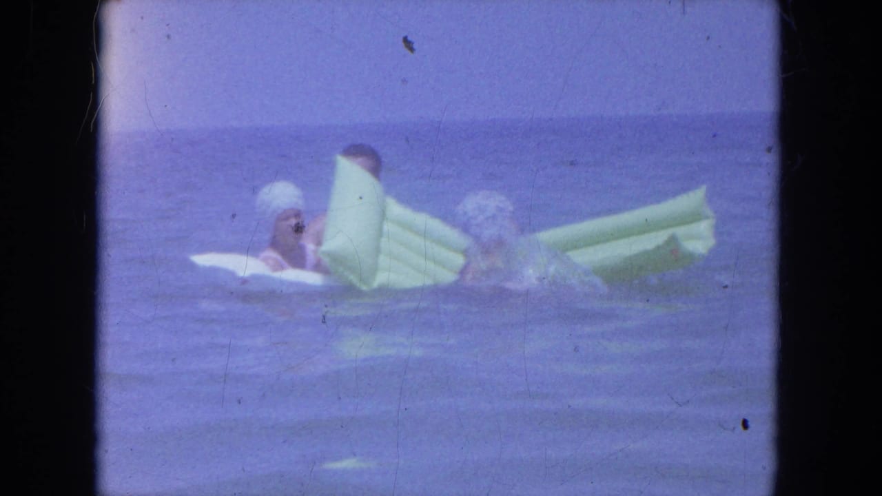 Two adults wearing swim caps sit partly upright with pale inflatable rafts in calm Clearwater ocean water.