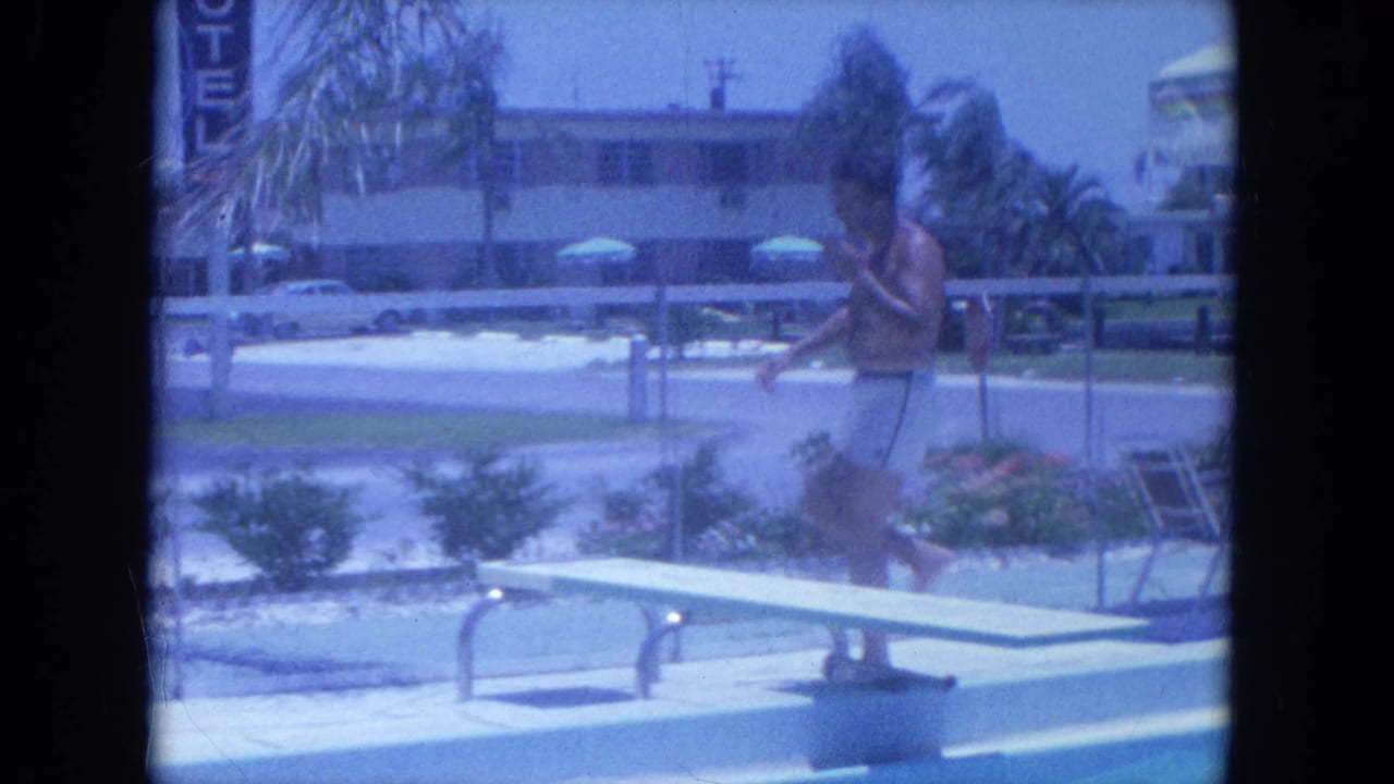 Man walking toward a poolside diving board near a motel sign and palm trees.