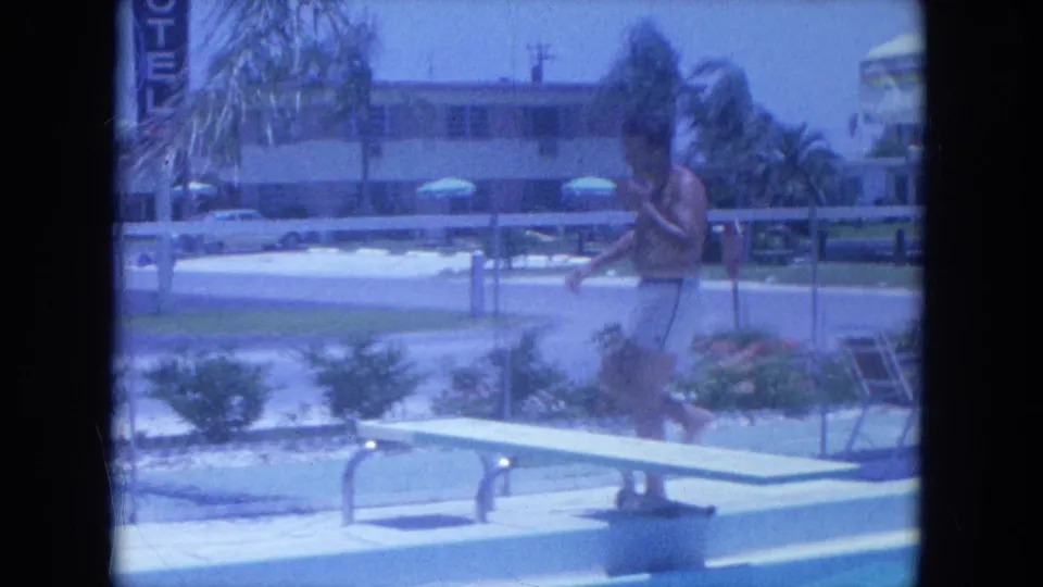 In 1964 Clearwater home-movie footage, a shirtless man approaches a motel pool diving board with palms and parked cars behind him.