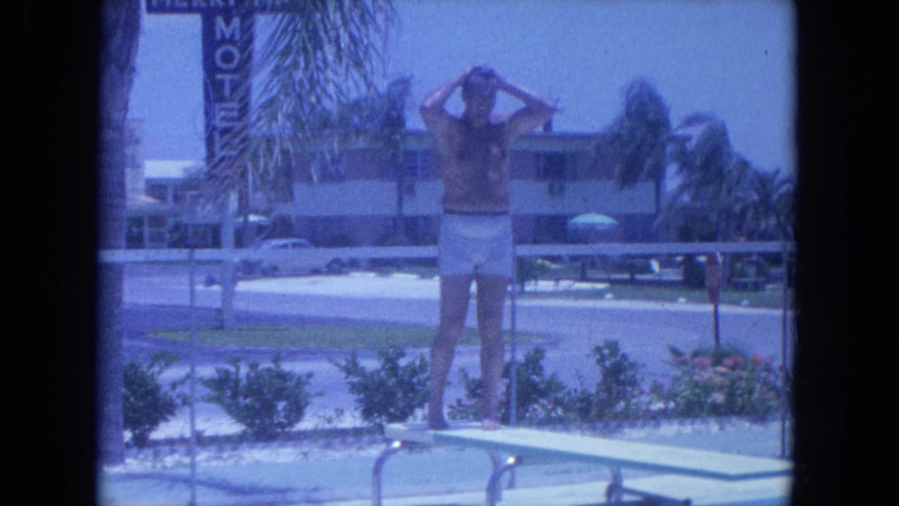 Man standing on a diving board with hands at his head near a motel pool.