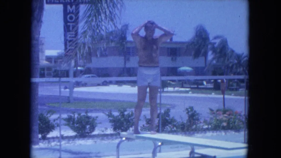 The man stands on the diving board at a Clearwater motel pool, with palms, signage and low-rise buildings in the background.