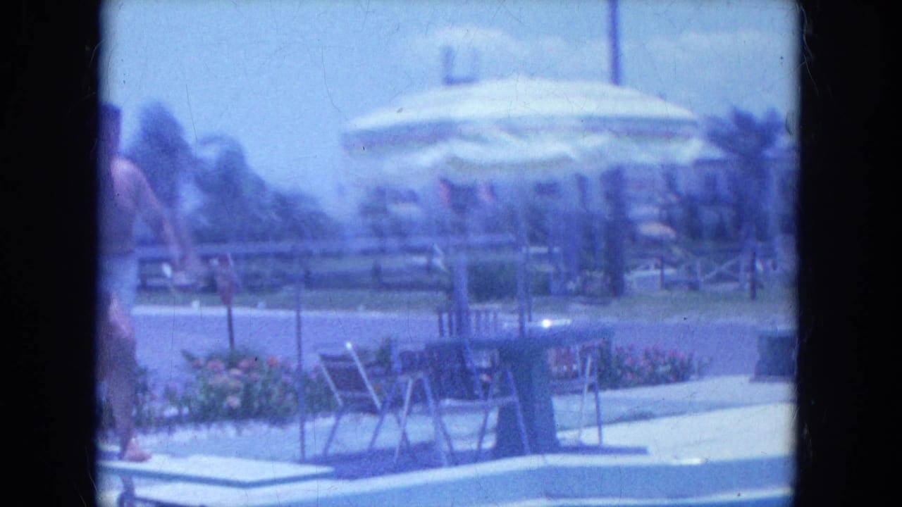 The motel pool area shows the diving board, lounge chairs, umbrellas, flower beds and people gathered near the deck.