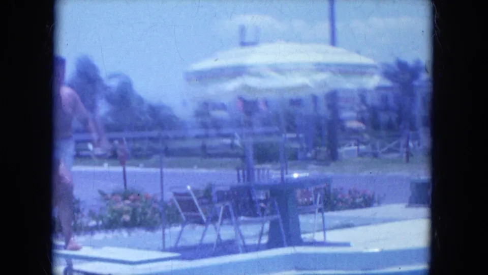 The motel pool area shows the diving board, lounge chairs, umbrellas, flower beds and people gathered near the deck.