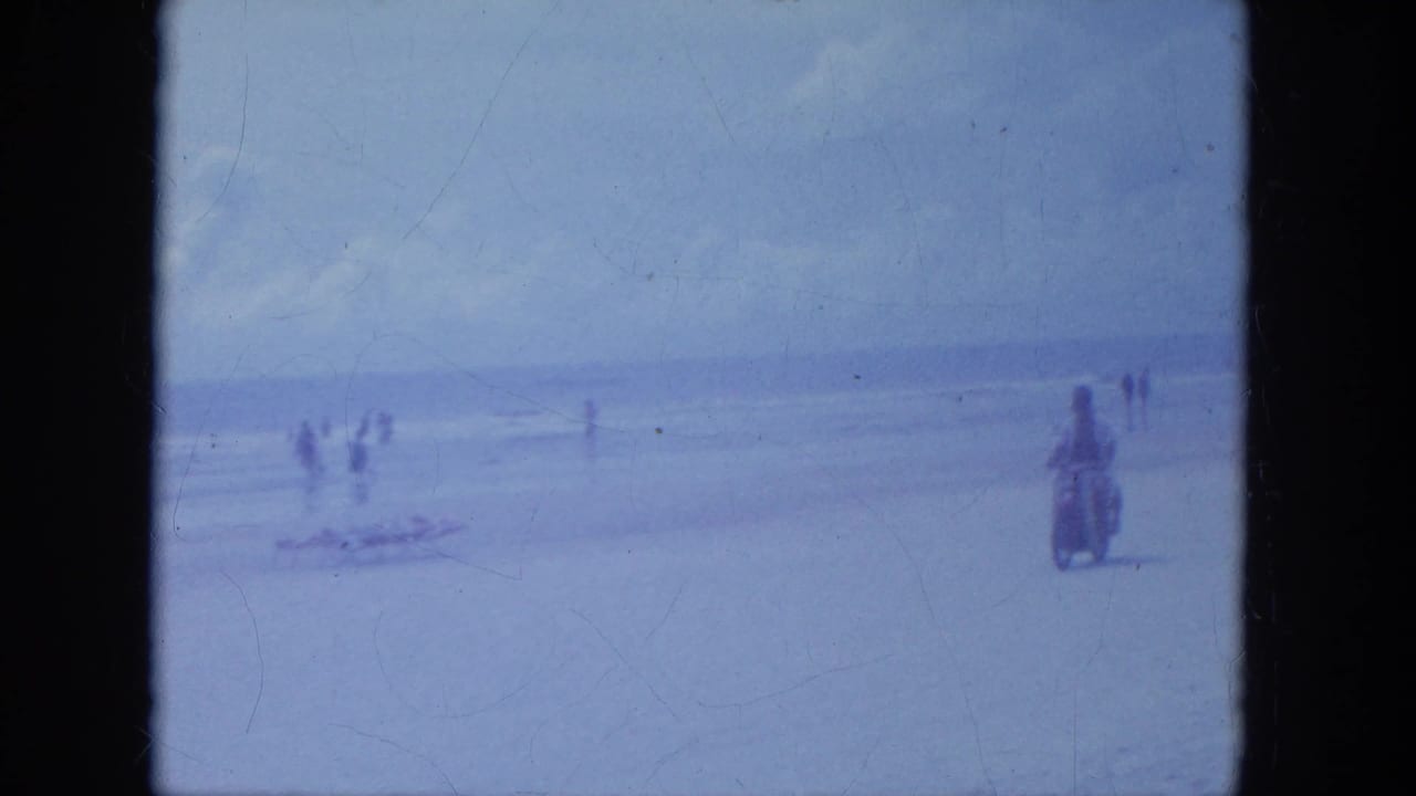 Two people ride a motorcycle across wet beach sand with bathers and surf in the distance.