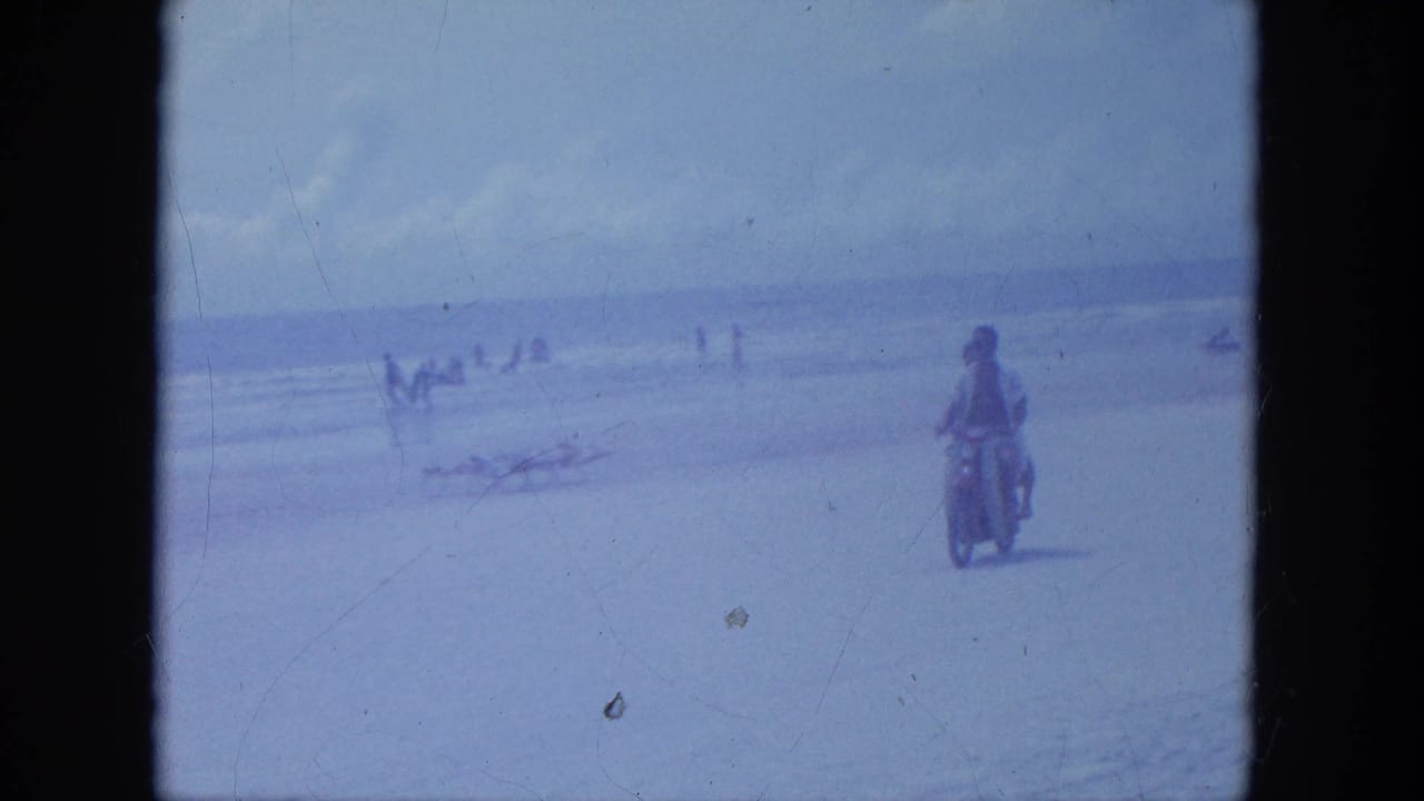 A motorcycle with two riders travels along the beach with small groups near the waterline.